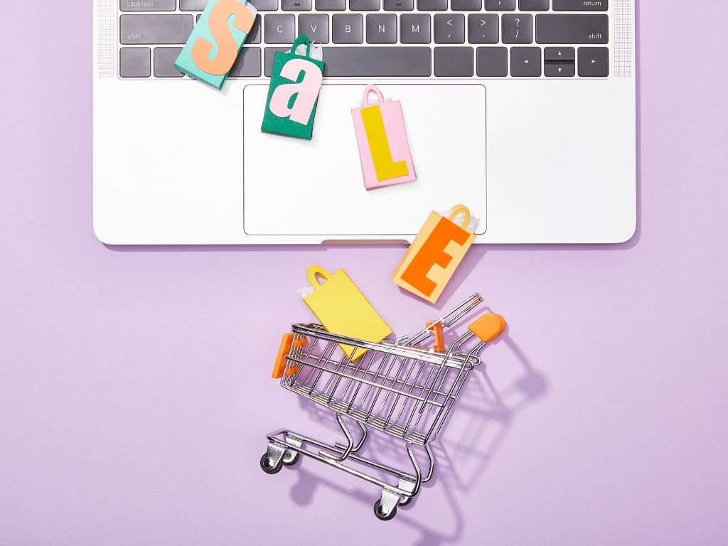 Símbolos de una experiencia de compra en línea: Letras "SALE", bolsas de compra pequeñas y un carrito de supermercado saliendo de un computador, representando la experiencia del cliente.