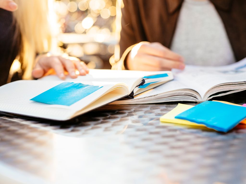 Primer plano de dos personas estudiando o trabajando al aire libre, con cuadernos abiertos y varias notas adhesivas de colores (azul y amarillo) sobre una mesa metálica, con la luz del sol de fondo.