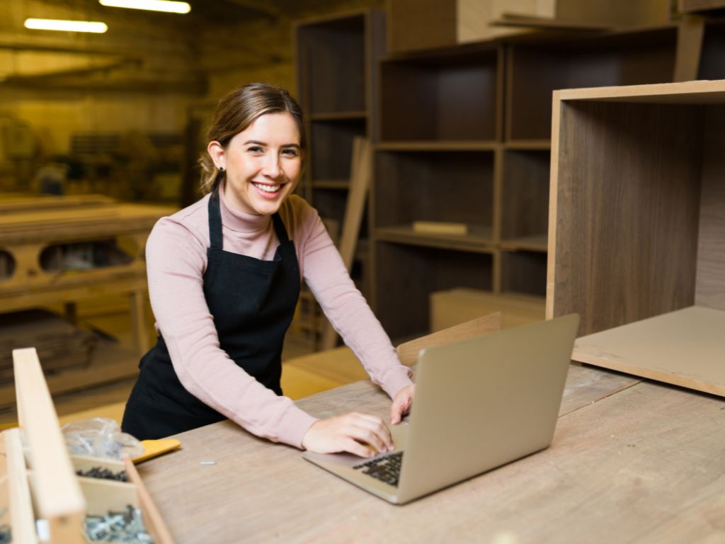 Joven dueña de negocio de carpintería sonriendo mientras usa un computador para gestionar pedidos y cotizaciones en su taller. Para ello, hace uso de la fibra óptica. 
