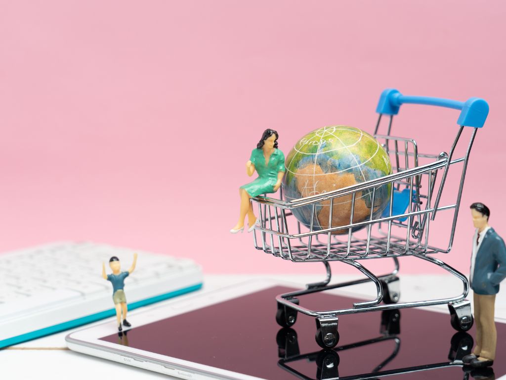 Dos pequeñas figuras, un hombre y una mujer, se sitúan alrededor de un carrito de compras en miniatura que contiene un globo terráqueo. La imagen representa el concepto de empresas en cyberday y el comercio electrónico.