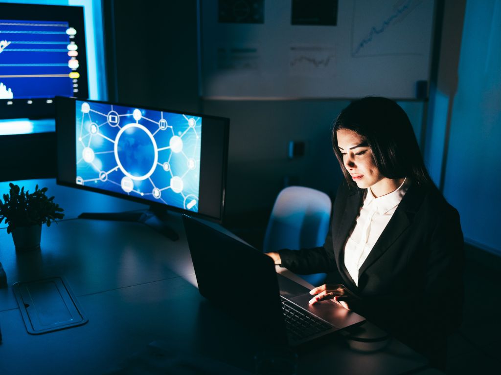 Una mujer concentrada trabaja en su computadora en una oficina oscura, con monitores que muestran gráficos y diagramas de análisis de datos.