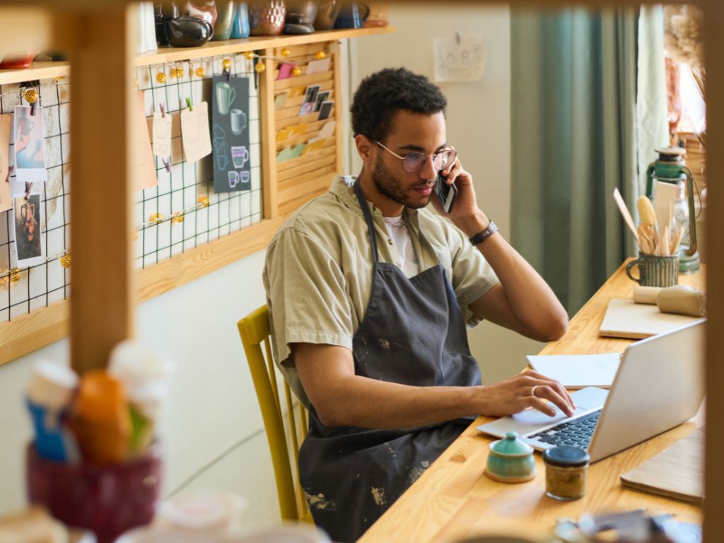 Joven emprendedor en su taller, está hablando por teléfono mientras investiga qué es el vpn en el celular para proteger su negocio digital.