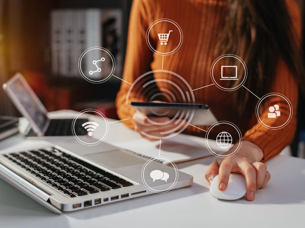Mujer usando un computador y una tablet, rodeada de iconos que representan el comercio electrónico, redes sociales, y comunicación. La imagen ilustra qué es una estrategia omnicanal. 