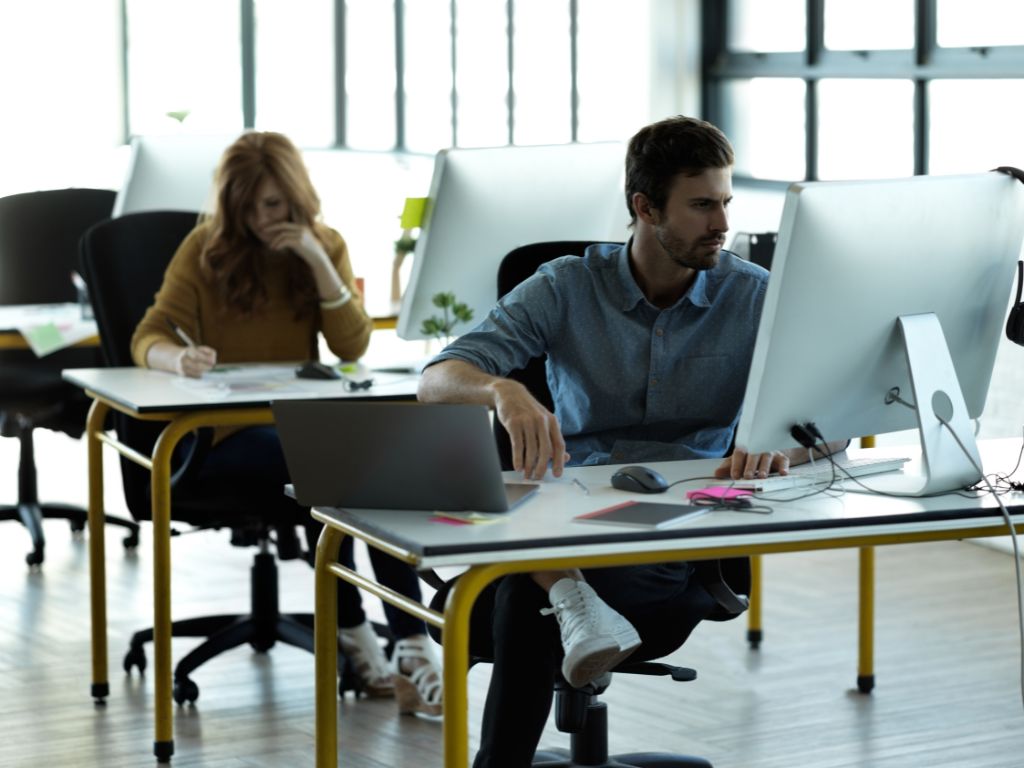 Un hombre dueño de una empresa, vestido con una camisa marrón, trabaja concentrado en su computador. Se encuentra sentado en un escritorio en una oficina moderna con paredes de cristal y reflejos.