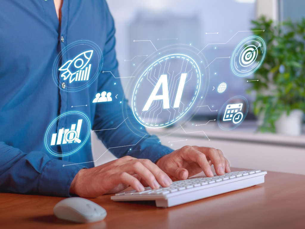 Hombre escribiendo en un teclado con superposiciones holográficas de inteligencia artificial, que incluyen los iconos de 'IA', un cohete, personas, un gráfico y un objetivo. La imagen es representativa sobre por qué es buena la inteligencia artificial.