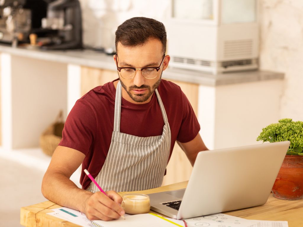Hombre joven con lentes y un delantal de rayas escribiendo en un cuaderno, mientras trabaja en un computador. La imagen representa el tema del contenido, la diferencia entre omnicanal y multicanal en un entorno de trabajo.